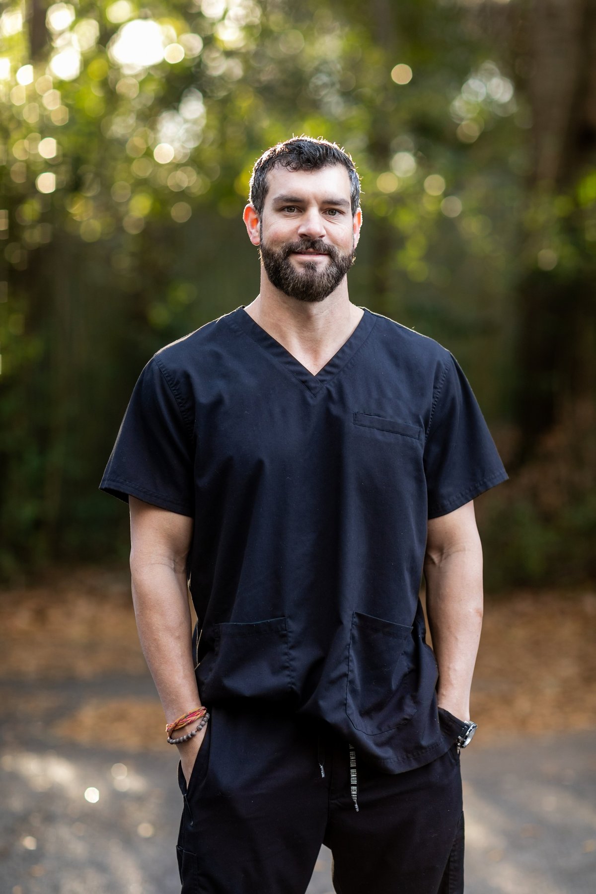 A doctor with dark hair and a beard in black scrubs standing outside in a natural green environment