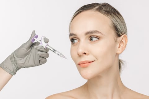 A woman confidently poses against a white background beside a syringe representing tear trough filler treatment